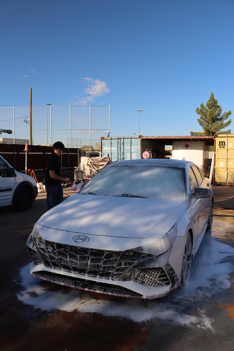 White luxury sedan being washed with soap and water at an outdoor car wash facility under clear blue sky