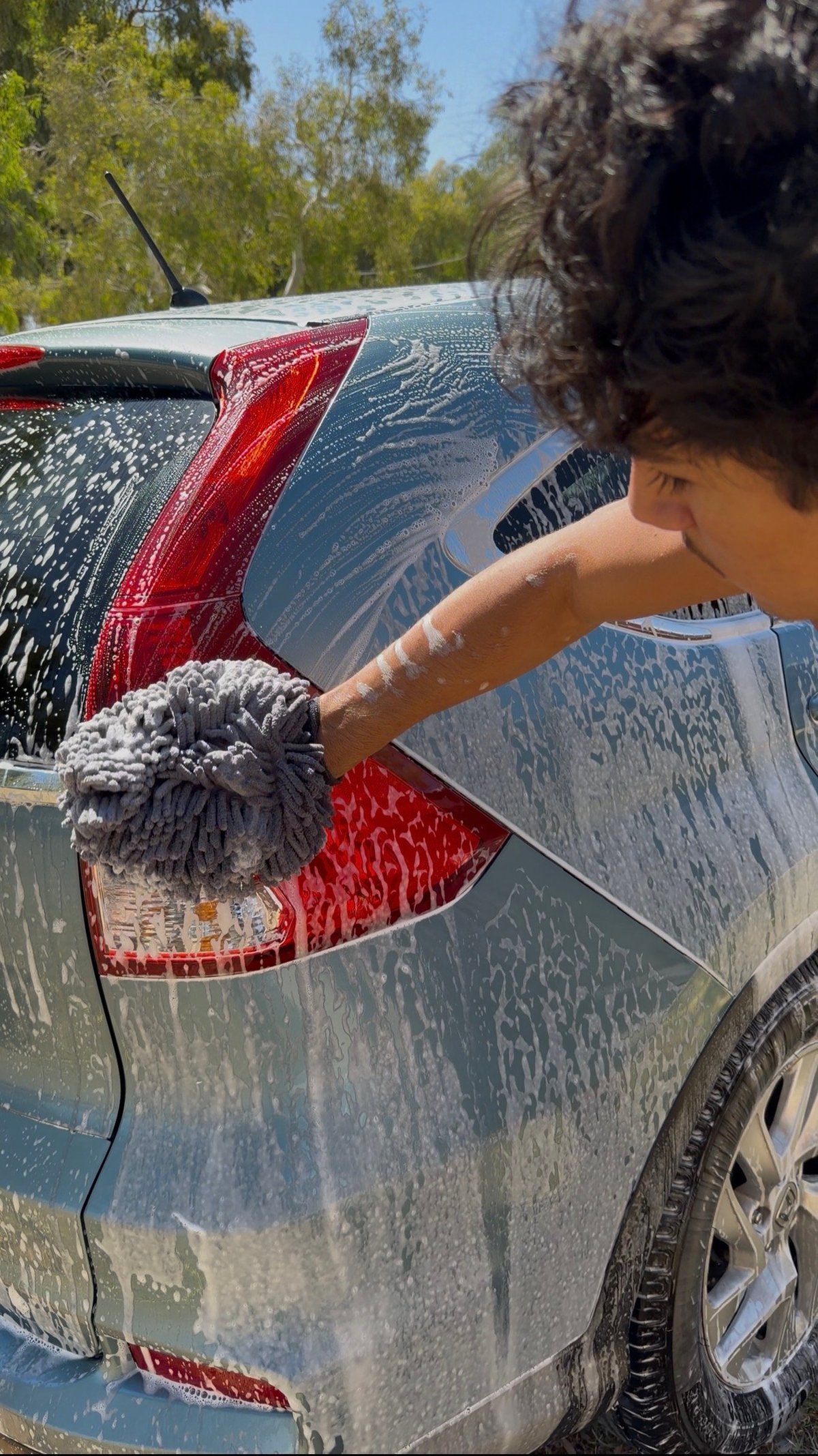Person washing a red and gray car with a soapy brush at an outdoor car wash on a sunny day